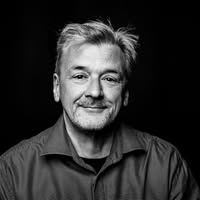 Schwarzweiß-Porträt eines Mannes mittleren Alters mit kurzen grauen Haaren, getragenem Hemd, freundlicher Ausstrahlung und dunklem Hintergrund.