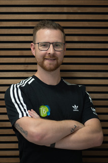 Mann mit Brille und Bart, trägt ein schwarzes Adidas-T-Shirt mit weißen Streifen, steht vor einer horizontale Holzlatwand.